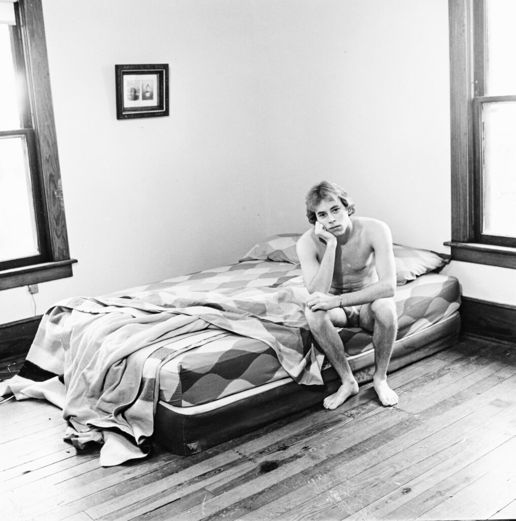 A young man sits in his underwear at the edge of a bed.