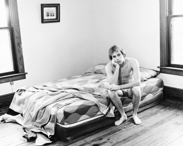 A young man sits in his underwear at the edge of a bed.